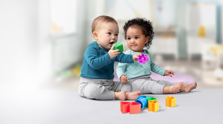 One baby observing as another infant places a green playing block into their mouth.