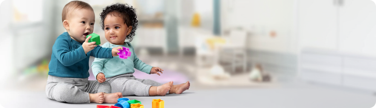 One baby observing as another infant places a green playing block into their mouth.